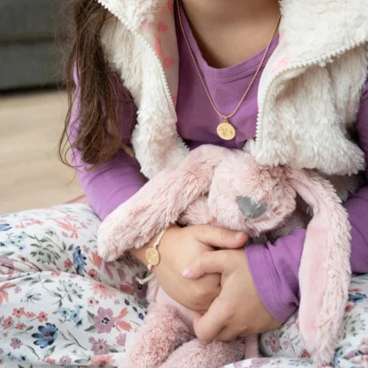 Kind met goudkleurige ketting Ik draag je en ronde vlinderhanger, met een roze knuffelkonijn op schoot.