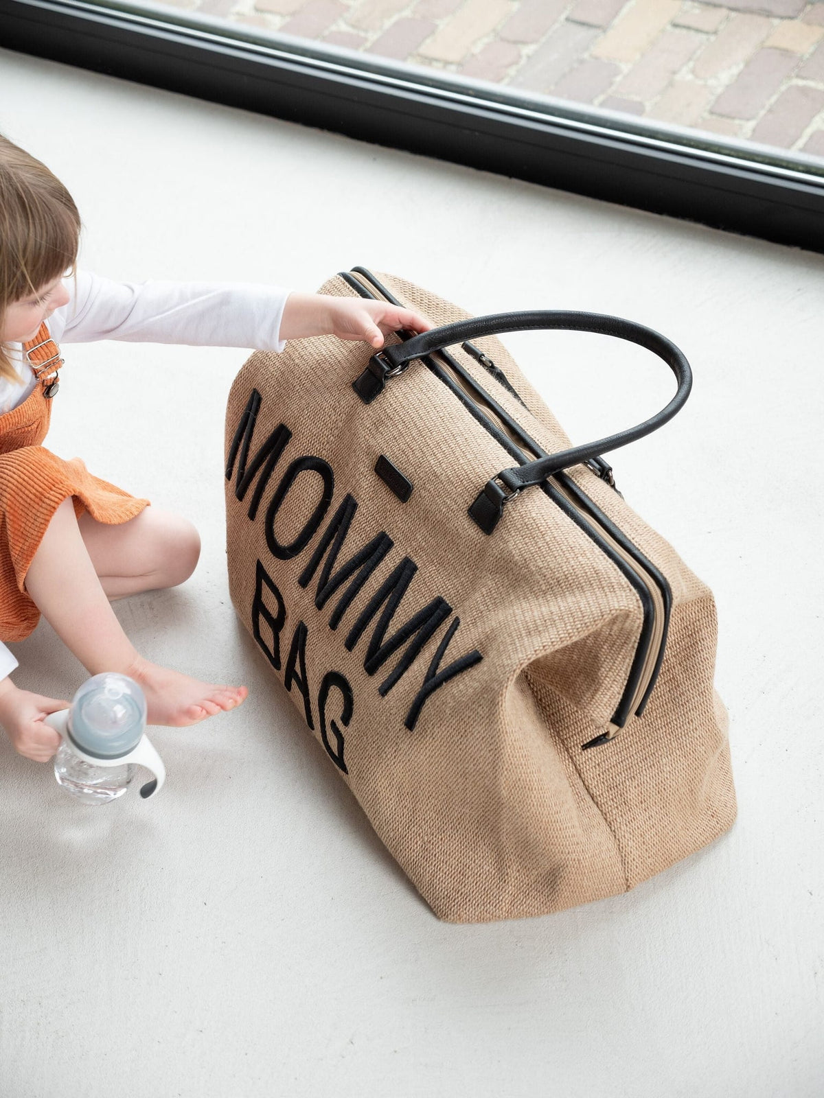 Beige Mommy Bag verzorgingstas met raffialook en zwarte handvatten naast een kind met een flesje.