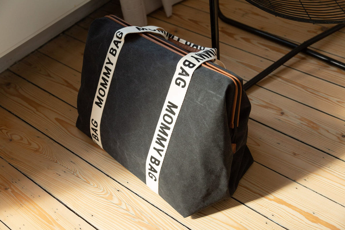 Zwarte Mommy Bag verzorgingstas met crèmekleurige draagbanden en bruine ritsdetails op een houten vloer.