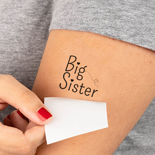 Big Sister-plaktattoo in zwarte letters met hartjes op een bovenarm, terwijl het witte papier wordt verwijderd