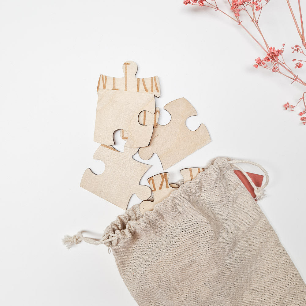 Houten puzzelstukjes met letters steken uit een beige linnen zakje, met roze droogbloemen op de achtergrond.