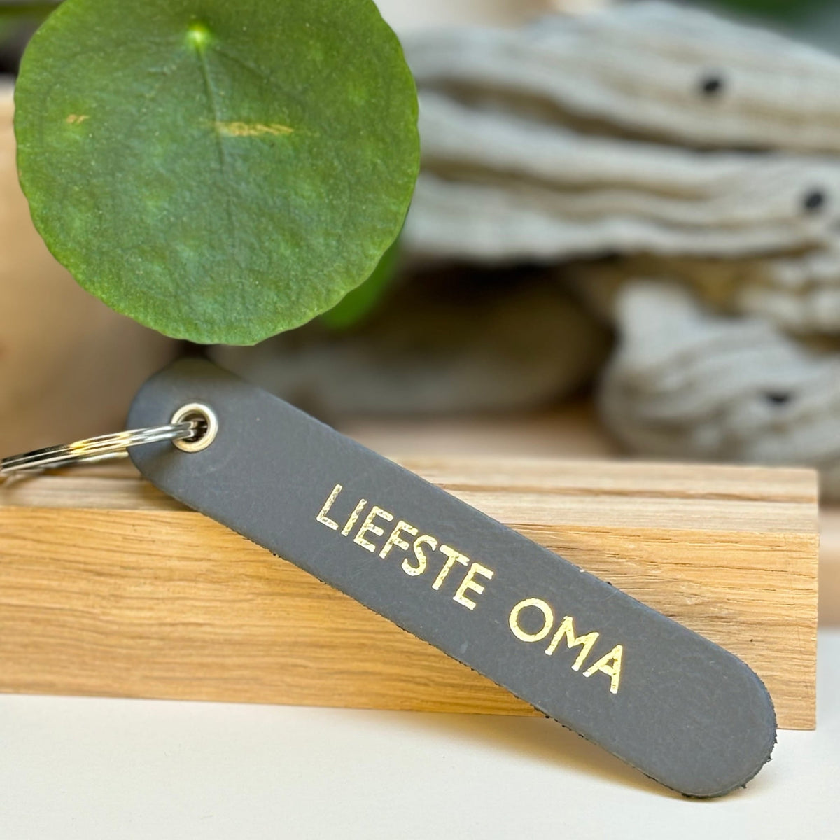 Grijze sleutelhanger met opdruk ‘Liefste Oma’ op een houten rand, met een groen blad op de achtergrond.