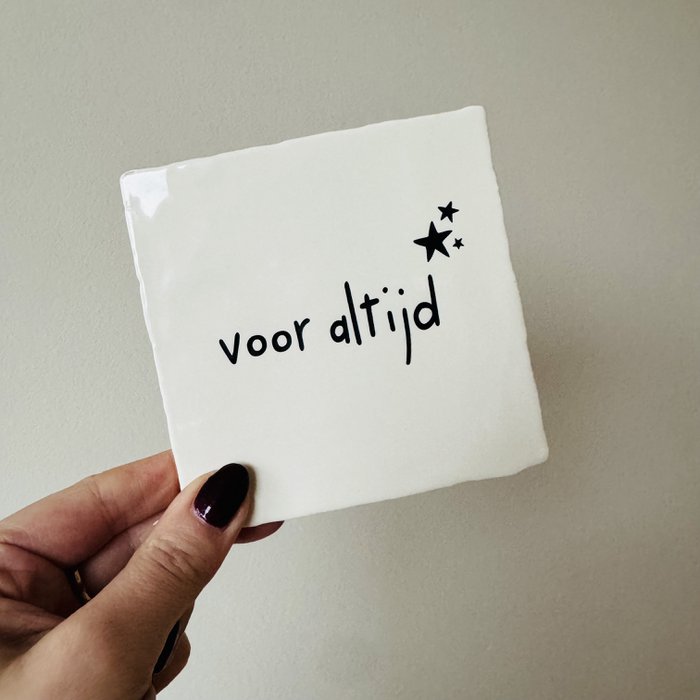Wit tegeltje met de zwarte tekst ‘voor altijd’ en drie zwarte sterren, vastgehouden in een hand.