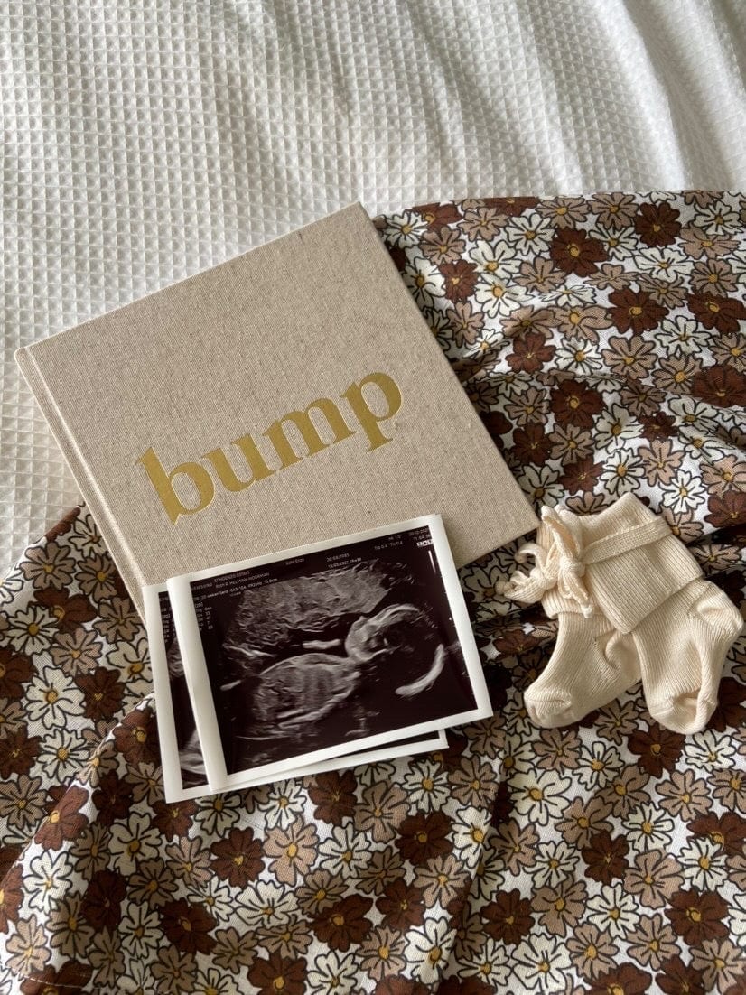 Bump zwangerschapsboek met nature kaft en gouden letters, naast echofoto's en babysokjes op een gebloemde ondergrond