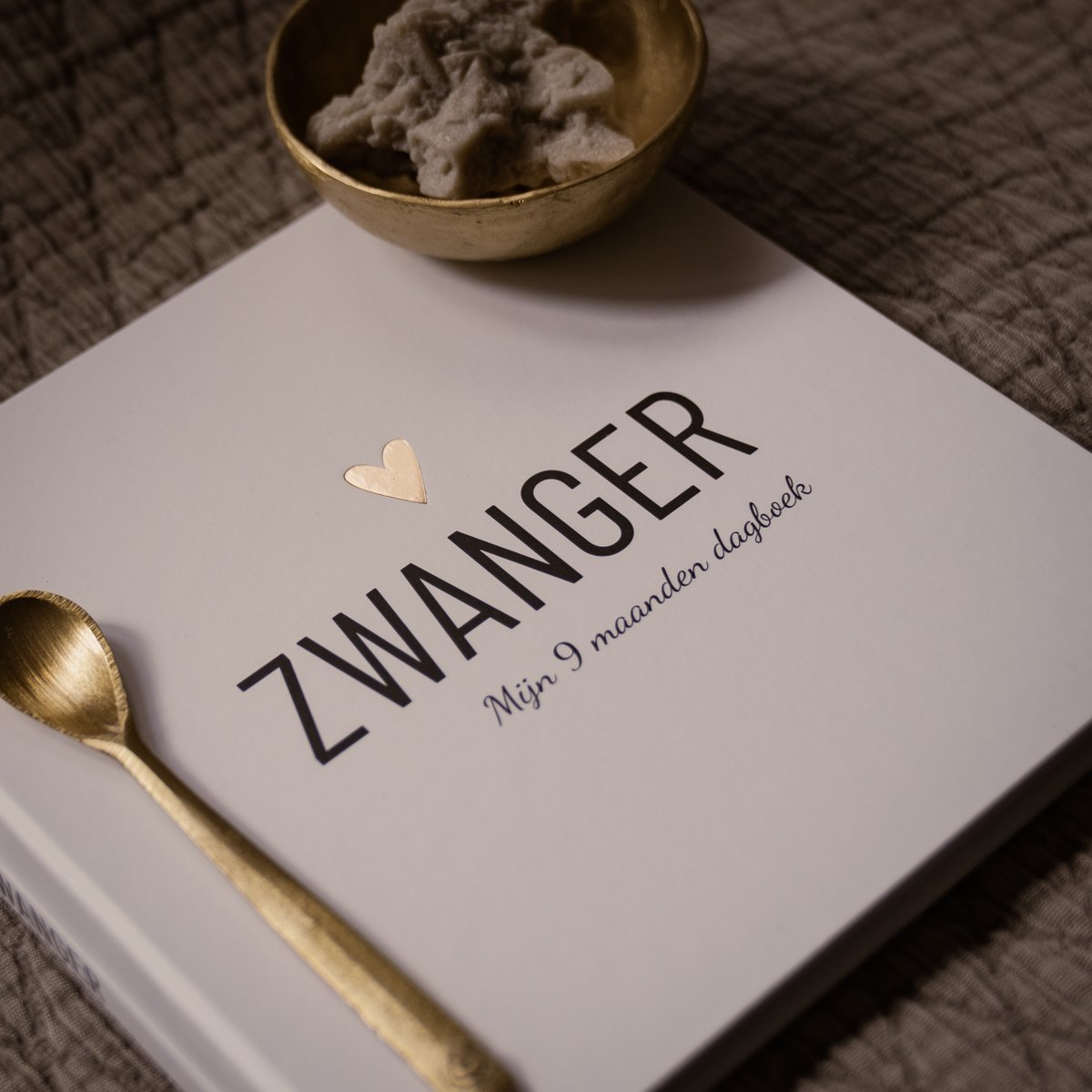 Schuin detail van het witte zwangerschapsdagboek met goudkleurig hartje, naast een lepel en schaaltje.