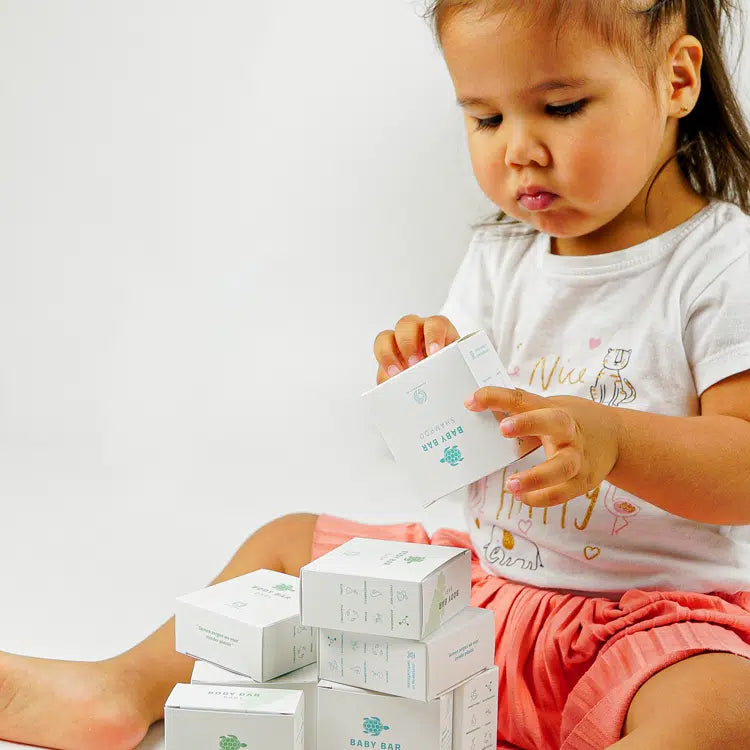 Kind bekijkt een doosje Shampoo Bar Baby met een stapel witte verpakkingen op schoot.