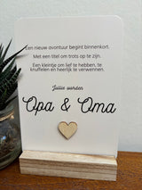 Witte A6 ansichtkaart met 'Jullie worden Opa & Oma' en een houten hartje, in een houten houder naast een plant.