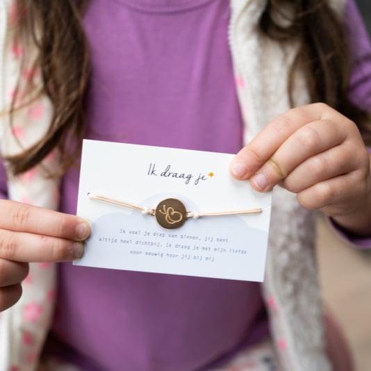 Kind houdt het kaartje met het gedicht Ik draag je en het lichtroze armbandje met goudkleurige vlinderbedel vast.