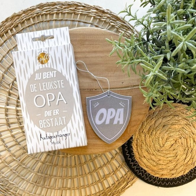 Schildvormige autogeurhanger met ‘Opa’ naast de verpakking op een houten plank, met een plant ernaast.
