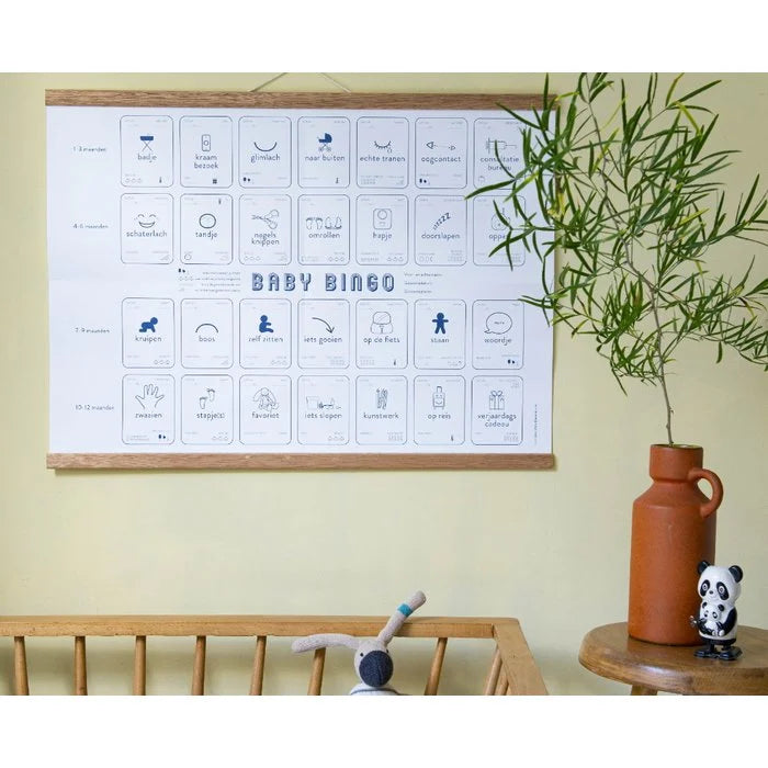 Baby Mijlpalen Bingo aan houten ophanglatten boven een babybed, naast een vaas met groene takken.