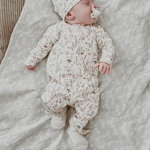 Baby met speen in boxpakje Dried Flowers met bijpassend mutsje en gebreide slofjes, liggend op een deken.