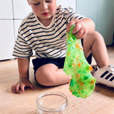 Een kind zit op de vloer en rekt groen Dino Dew-slijm met bedeltjes uit boven een open pot.