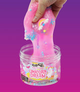 Een hand trekt roze Unicorn Dream-slijm met regenboogbedeltjes en gekleurde bolletjes uit de pot.