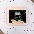 Houten echolijst met echofoto en de tekst 'It's a Boy' op een witte ondergrond met donkere stippen.