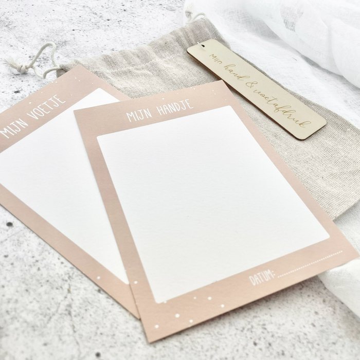 Twee beige kaartjes voor hand- en voetafdrukken met witte afdrukvlakken, naast het linnen bewaarzakje.