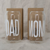 Twee hoge, transparante drinkglazen met witte teksten ‘Dad’ en ‘Mom’, voor kartonnen verpakkingen.