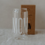 Hoog transparant drinkglas met wit opschrift ‘Mom’, met een bruine doos op de achtergrond.