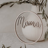 Lichtbruine houten krans met het woord mama leunt tegen een witte vaas met groene takken.