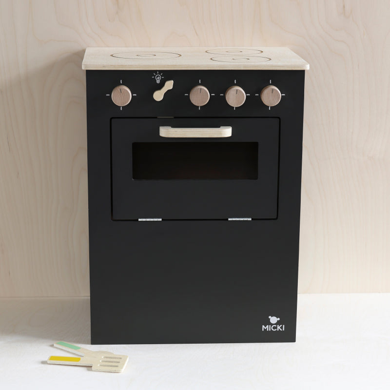 Zwarte houten speelkeuken tegen een houten wand, met speelgoedkeukengerei op de vloer.