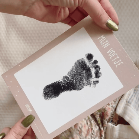 Kaart 'Mijn Voetje' met zwarte babyvoetafdruk en een lichtbruine rand, vastgehouden met twee handen.