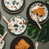Ronde kartonnen dinobordjes met groene geschulpte rand en dinosaurusprint, op een gedekte tafel met wafels en bestek.