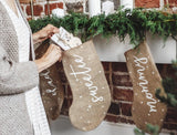 Persoon vult een jute kerstsok met een cadeau bij een haard met dennengroen en meerdere kerstsokken.