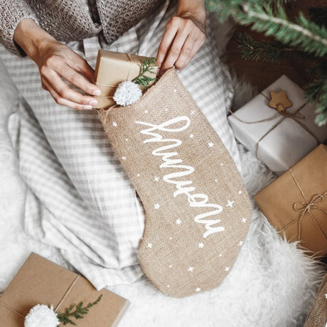 Jute kerstsok met witte tekst 'mommy' en sterretjes, gevuld met een cadeautje naast de kerstboom.