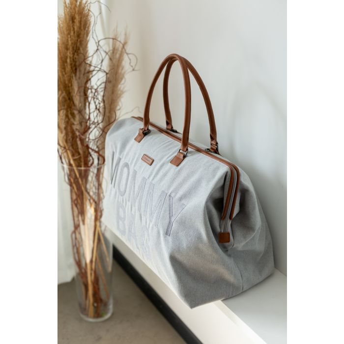 Grijze Mommy Bag verzorgingstas schuin van opzij op een witte plank naast een vaas met droogtakken.