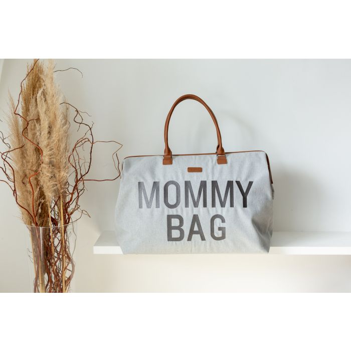Grijze Mommy Bag verzorgingstas met bruine handvatten op een witte plank naast droogbloemen.