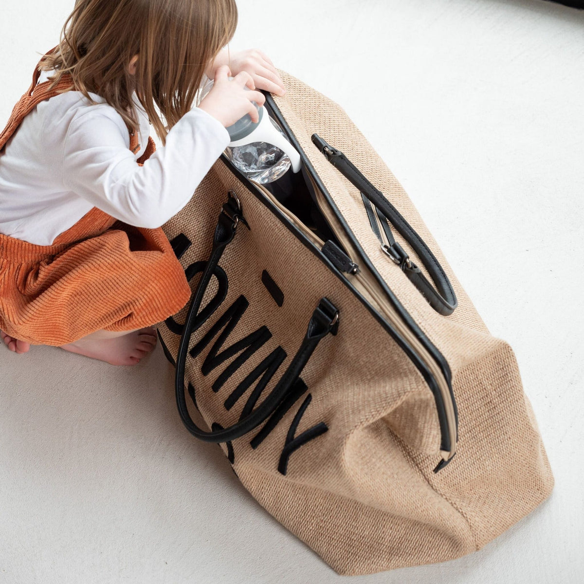 Een kind stopt een flesje in de geopende beige Mommy Bag verzorgingstas met raffialook.