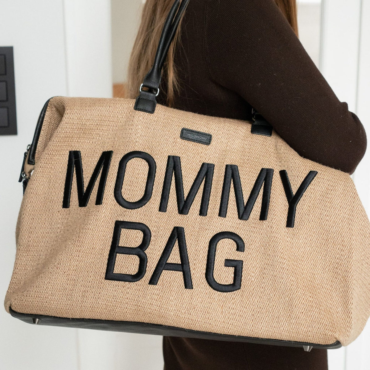 Close-up van de beige Mommy Bag verzorgingstas met zwarte letters en handvatten, gedragen over de schouder.