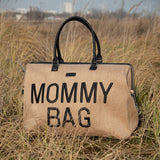 Beige Mommy Bag verzorgingstas met raffialook, zwarte handvatten en zwarte letters, buiten in het hoge gras.
