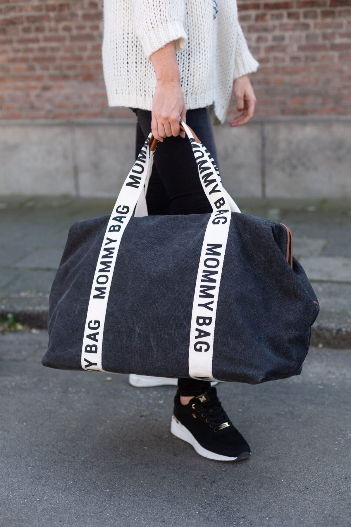 Persoon draagt een zwarte Mommy Bag verzorgingstas aan de crèmekleurige handgrepen op straat.