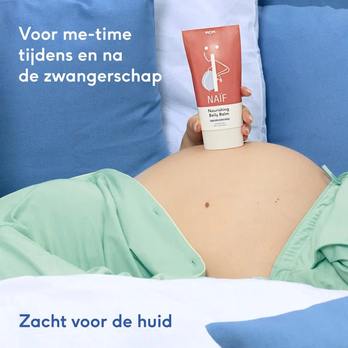 Zwangere vrouw houdt een rood-witte tube Naïf Nourishing Belly Balm boven haar blote buik, tussen blauwe kussens.