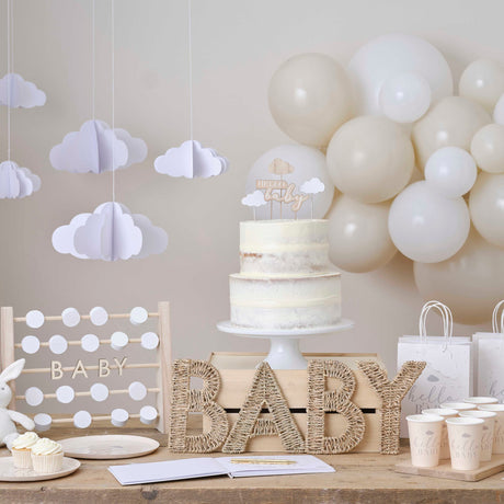 Naturelkleurige rieten letters ‘baby’ op een feesttafel met taart, witte ballonnen en hangende wolkjes.