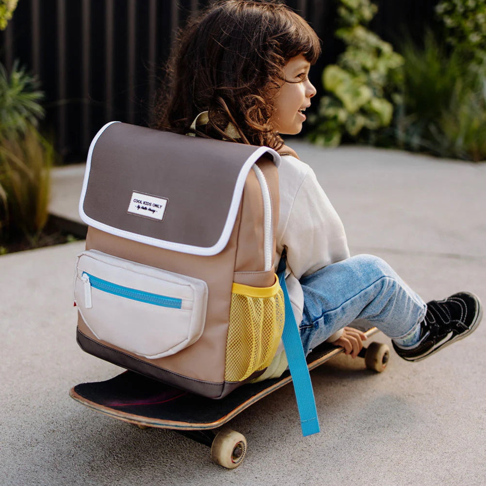 Kind met de Mini Walnut-rugzak op de rug, zittend op een skateboard buiten.
