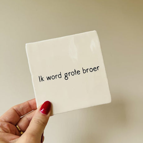 Wit tegeltje met de zwarte tekst 'Ik word grote broer', vastgehouden in een hand tegen een beige achtergrond.