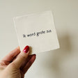 Wit vierkant tegeltje met de zwarte tekst 'Ik word grote zus', vastgehouden in een hand tegen een lichte muur.