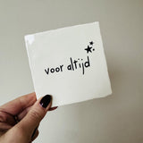 Wit tegeltje met de zwarte tekst ‘voor altijd’ en drie zwarte sterren, vastgehouden in een hand.
