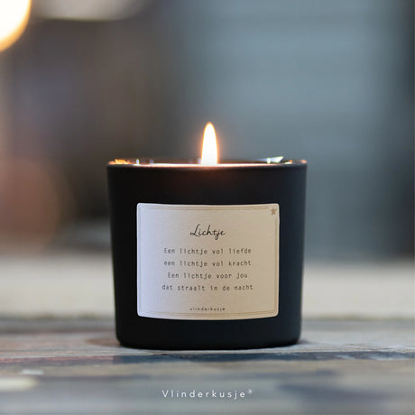 Brandende geurkaars Lichtje in een zwart glas met een wit etiket met gedichtje, op een houten ondergrond.