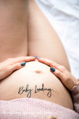 Zwarte plaktattoo 'Baby Loading...' op een zwangere buik, met twee handen rond de buik en een roze trui.