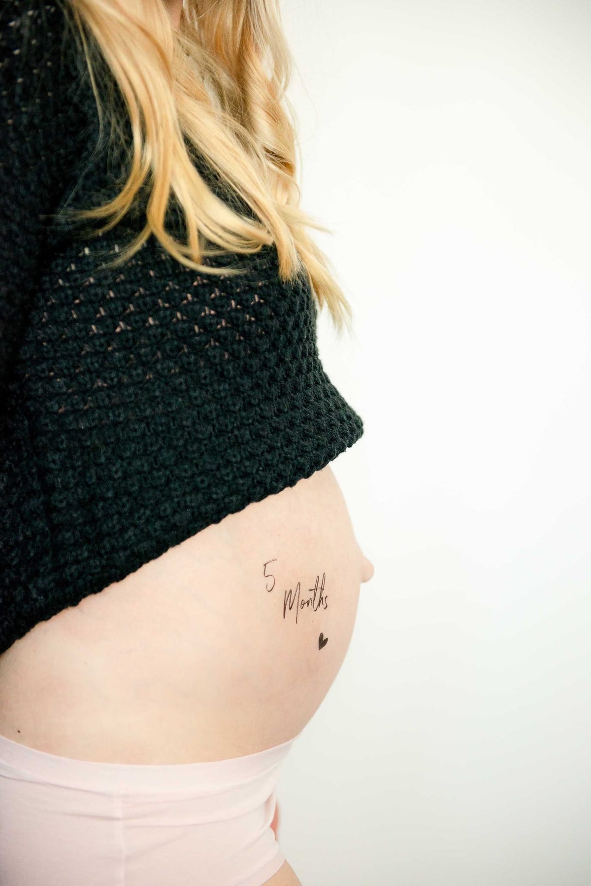 Plaktattoo '5 Months' met een hartje op een zwangere buik, onder een zwarte trui en boven een lichtroze slip.