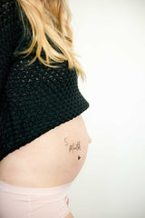 Plaktattoo '5 Months' met een hartje op een zwangere buik, onder een zwarte trui en boven een lichtroze slip.