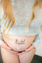 Zwarte plaktattoo met de tekst ‘It's a Girl’ op een zwangere buik onder een grijze top.