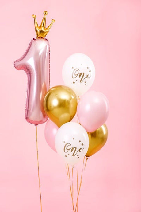 Roze, gouden en witte ballonnen met ‘One’-print naast een roze cijferballon 1 met gouden kroon, tegen een roze achtergrond.