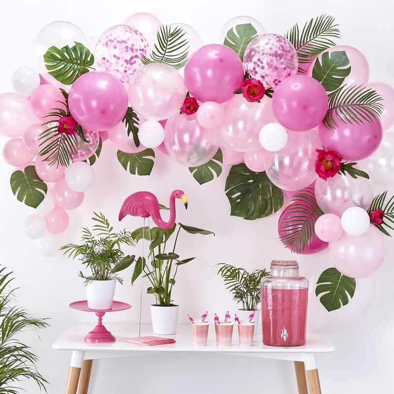 Roze ballonnenboog met confettiballonnen, groene bladeren en bloemen boven een feesttafel met flamingodecoratie