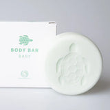 Lichtgroene Body Bar Baby met schildpadmotief naast een witte verpakking op een lichtgrijze achtergrond.