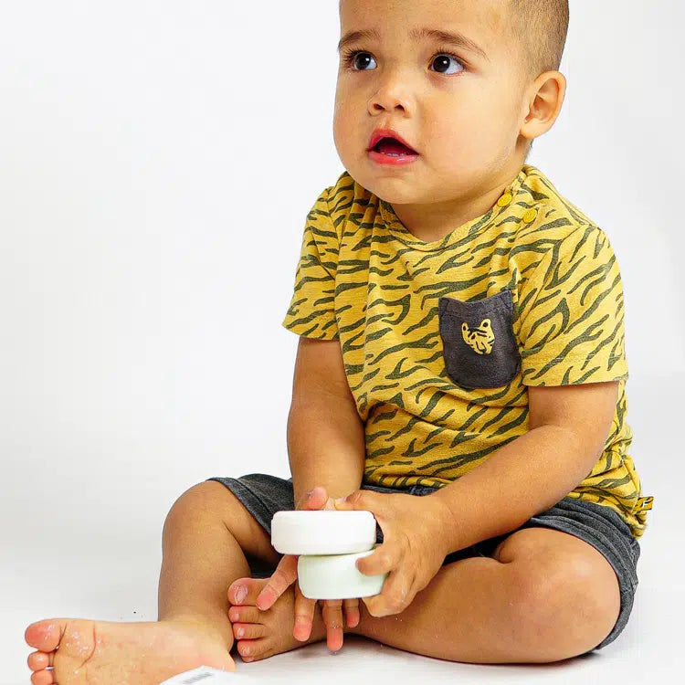 Zittend kind in een geel shirt met tijgerstrepen houdt twee ronde zeepbars vast, waaronder de lichtgroene Body Bar Baby.