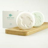 Lichtgroene Body Bar Baby achter een witte bar, beide met schildpadmotief, op een houten plankje naast verpakkingen.