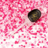 Transparante Confetti Push Pop met zwart etiket, liggend tussen roze en witte ronde confetti.
